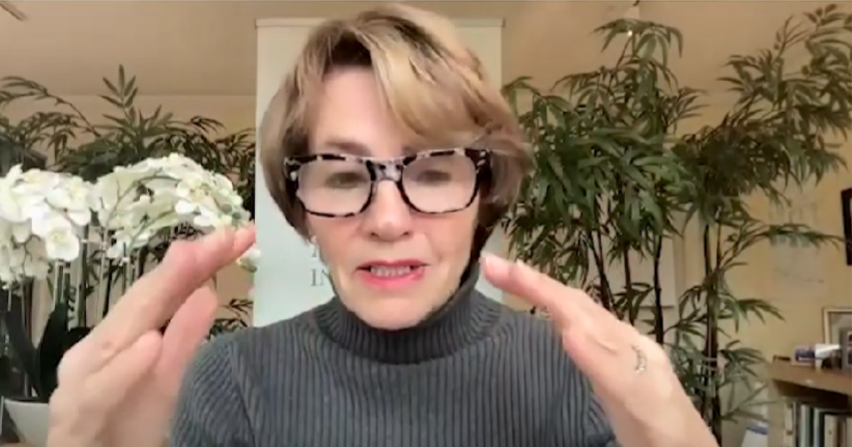 A woman gestures at the camera. She has short, light brown hair, a gray turtleneck, and trendy speckled glasses. Behind her are a number of plants.